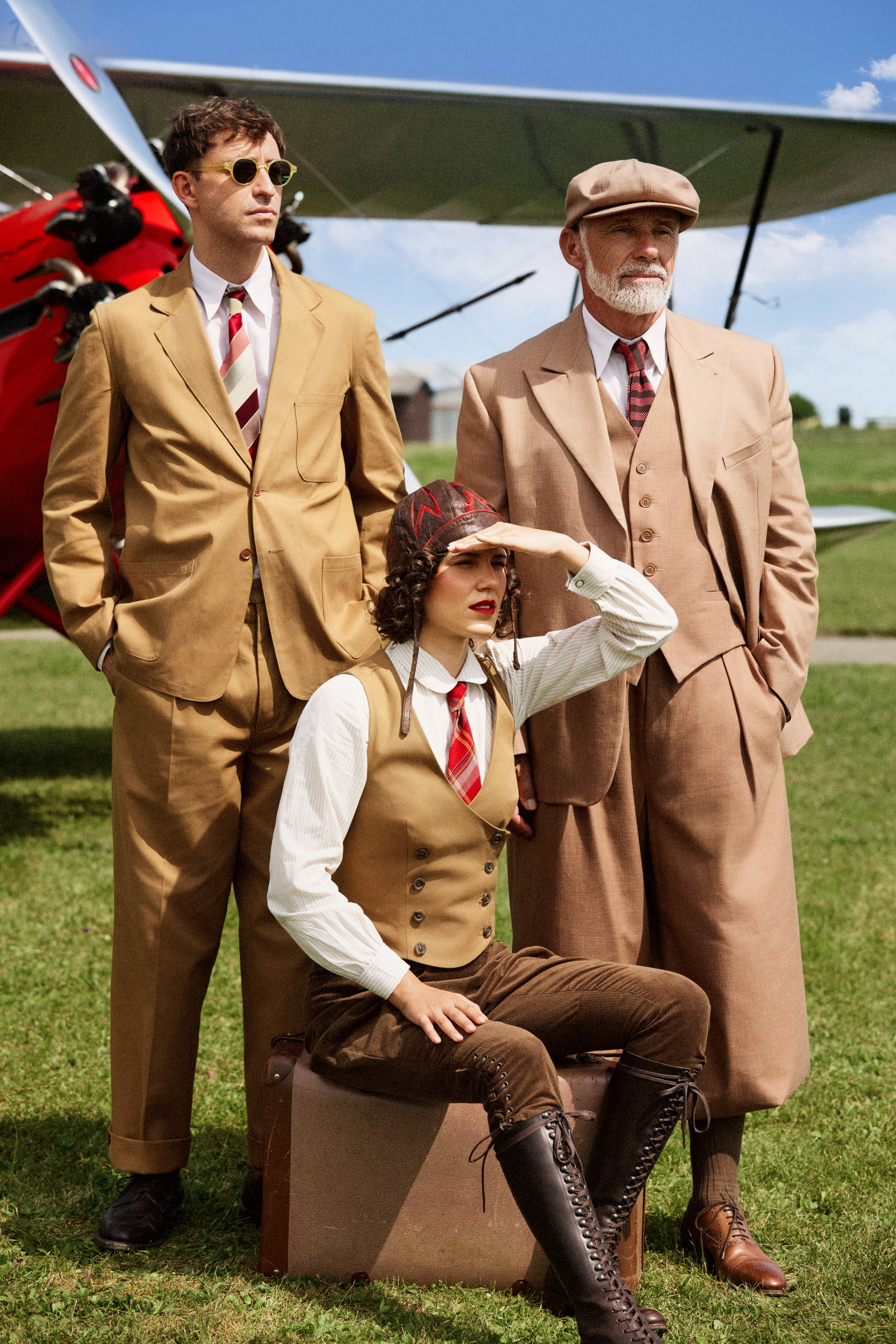 Authentische 1930er Mode von Vecona Vintage: Oldtimer-Treffen oder Tweed Run inspirierter Look mit beigem Herrenanzug, Knickerbockern und braunen Reitstiefeln, posierend auf einem Flugfeld vor einem historischen Flugzeug. Mehr zu Vintage Fashion auf unser