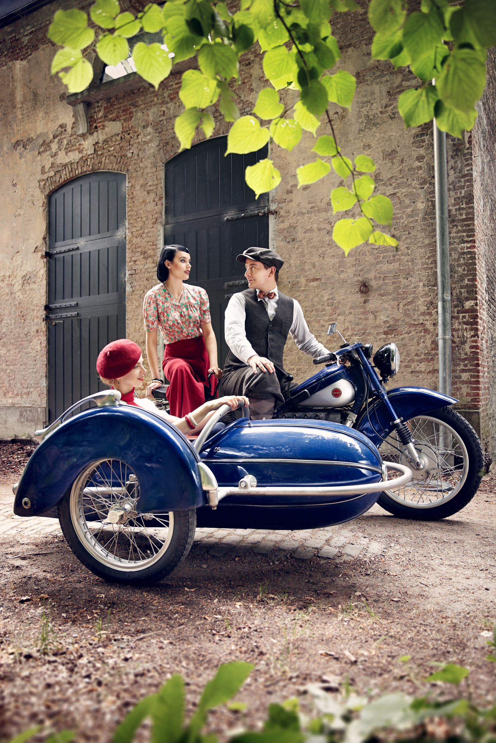 Vecona Vintage Motorrad-Look: Drei Personen in authentischer Vintage Mode der späten 1930er und 1940er Jahre, sitzend auf einem blauen Oldtimer-Motorrad mit Beiwagen vor einem historischen Gebäude. Perfekte Kleidung für Motorrad-Treffen und Rockabilly Sty